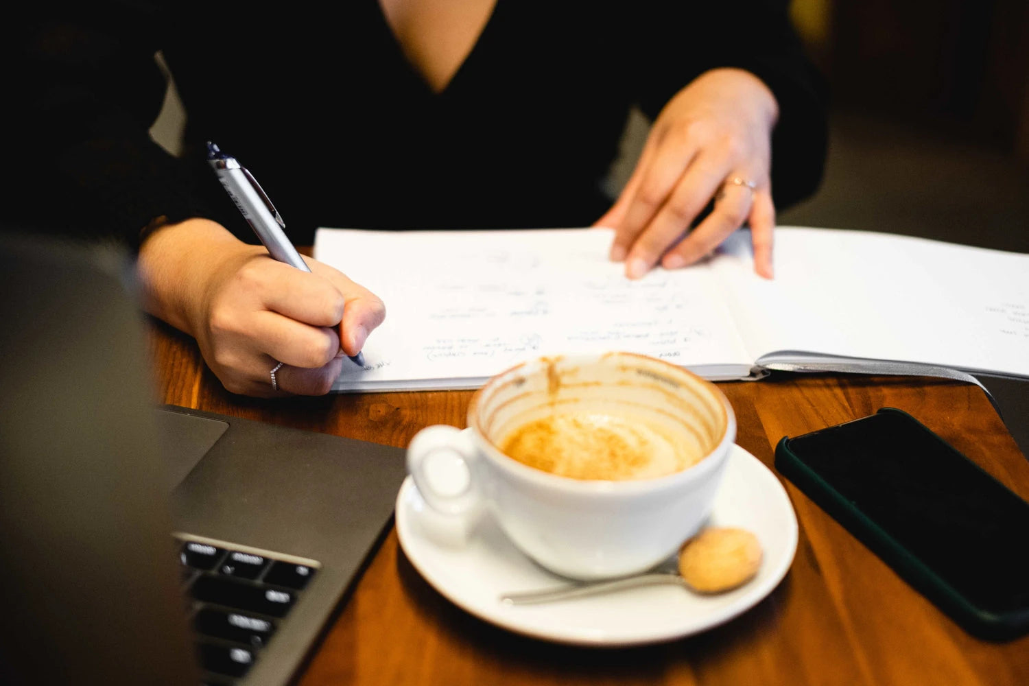 Person writing on a notepad with a cup of coffee and smartphone on a table.